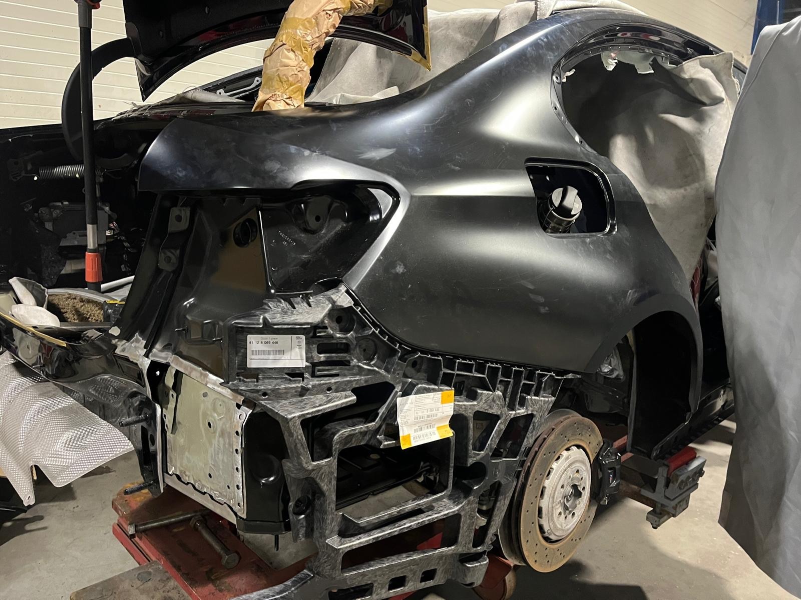 Czarny samochód w trakcie naprawy blacharskiej. Widoczne elementy karoserii w surowym stanie, przygotowane do dalszej obróbki. Detale mocowań i fragment tarczy hamulcowej.