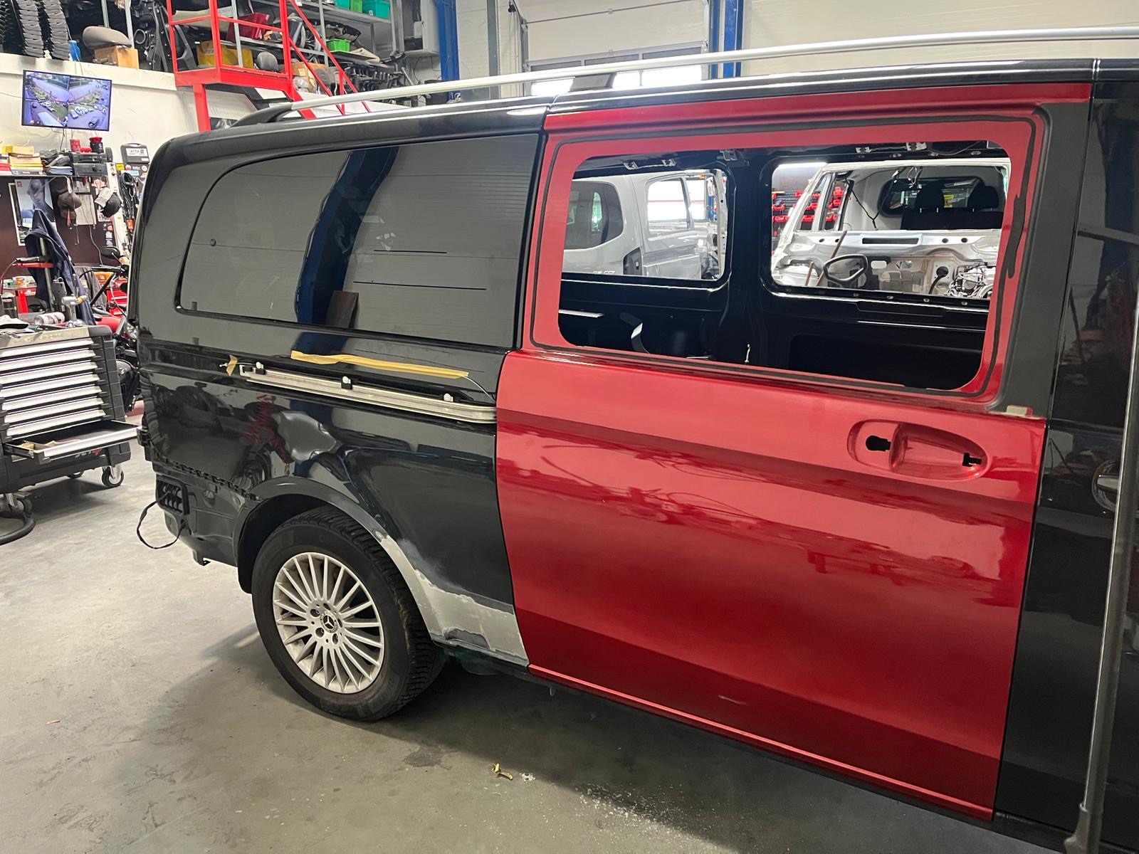 Częściowo zdemontowany, czarno-czerwony van w warsztacie. Widoczne ślady napraw blacharskich i przygotowania do lakierowania. W tle inne pojazdy w trakcie napraw.