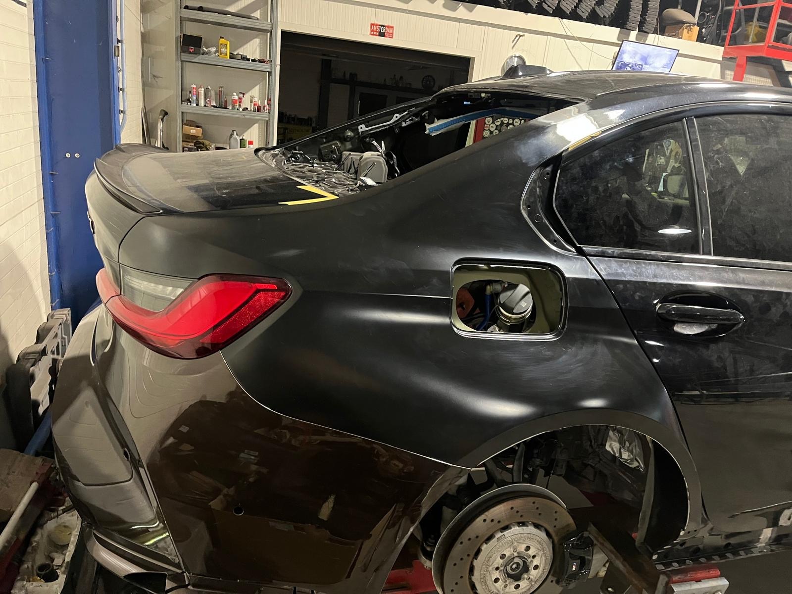 Czarny samochód w trakcie naprawy blacharsko-lakierniczej, widoczne elementy karoserii przygotowane do lakierowania, brak koła, warsztatowe otoczenie.