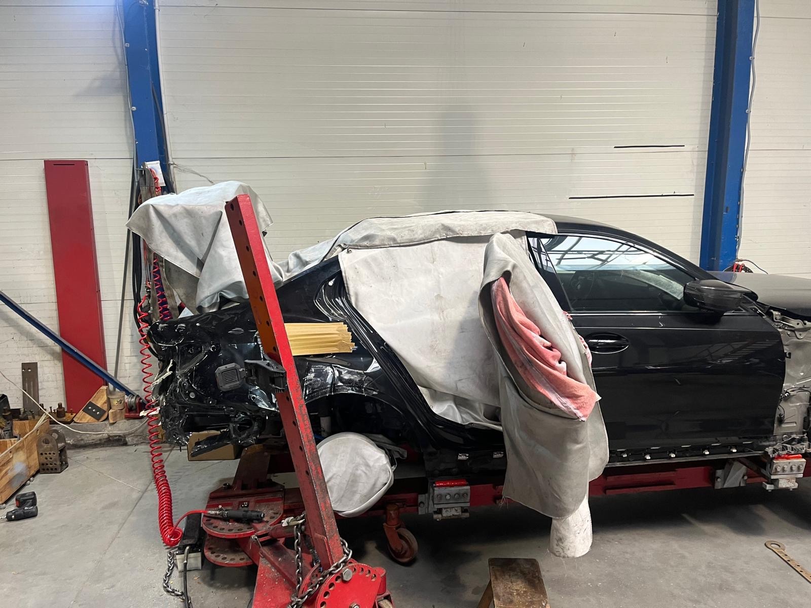 Czarny samochód na czerwonej ramie w warsztacie blacharskim, z uszkodzonym tyłem i częściowo zakryty płachtą. Widoczne narzędzia i elementy wyposażenia warsztatu.