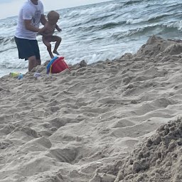 DAN-BRUK Daniel Palmowski - Radosny moment na plaży: mężczyzna podrzuca dziecko na tle falującego morza, obok kolorowe zabawki w piasku. Scena pełna beztroski i letniej zabawy.