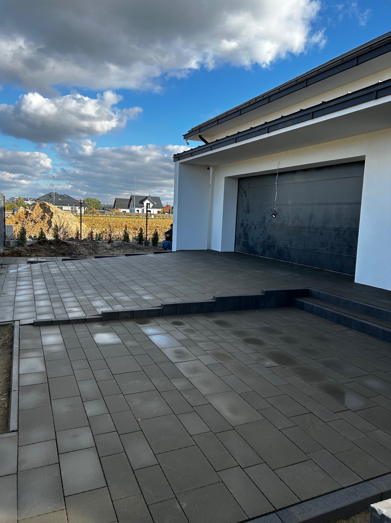 Ułożona, grafitowa kostka brukowa przed nowoczesnym domem z garażem. Widoczne schody z kostki i przygotowany teren pod nasadzenia. Błękitne niebo z chmurami w tle.