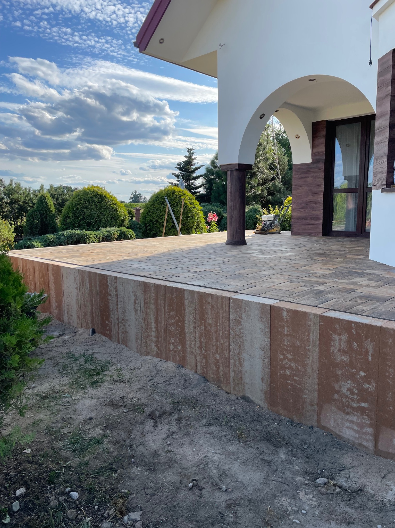 Wykończony taras z kostki brukowej w odcieniach brązu i szarości, z widoczną kolumną podtrzymującą zadaszenie i zielenią w tle. Widok na dom z łukowym wejściem.