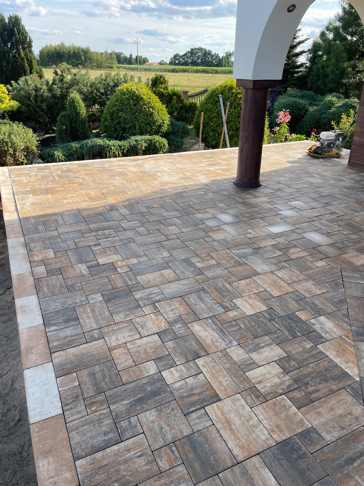 Ułożona kostka brukowa w odcieniach brązu i szarości na tarasie z kolumną. W tle zielony ogród i błękitne niebo. Widoczna zagęszczarka.