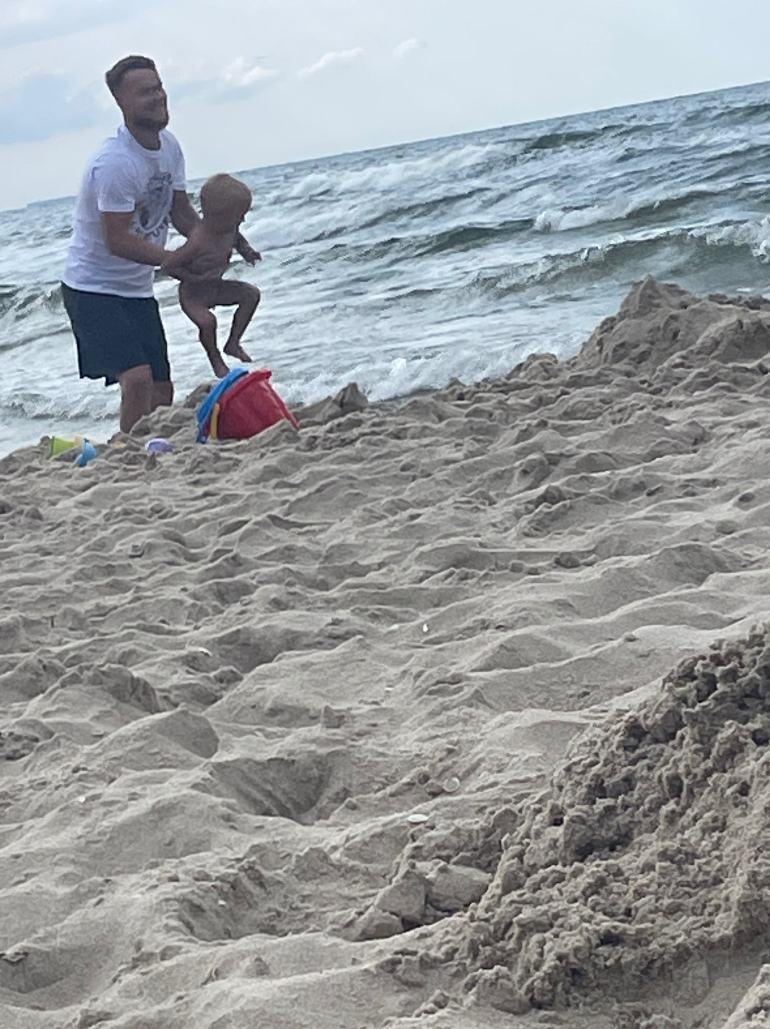 Radosny moment na plaży: mężczyzna podrzuca dziecko na tle falującego morza, obok kolorowe zabawki w piasku. Scena pełna beztroski i letniej zabawy.