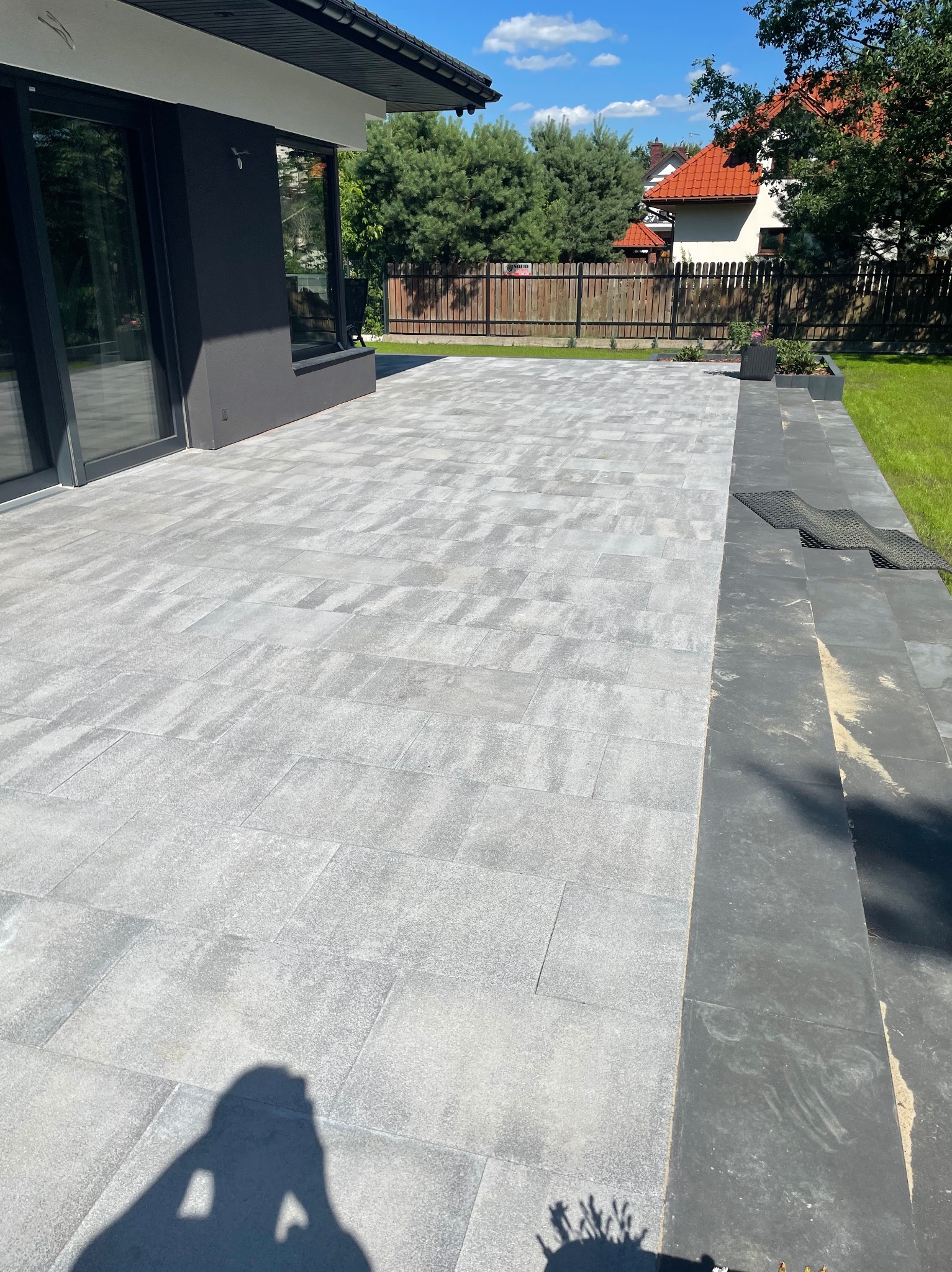 Nowoczesny taras z szarej kostki brukowej, przylegający do domu z dużymi oknami. Widoczne elementy zieleni i ogrodzenia w tle. Minimalistyczny design.