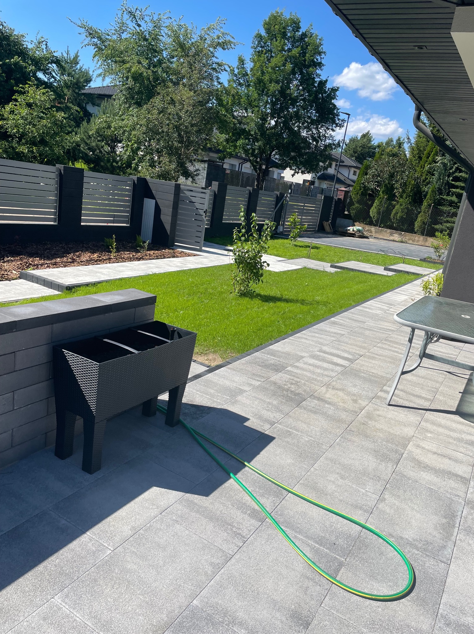 Nowoczesny ogród z szarą kostką brukową, zielonym trawnikiem i minimalistycznym ogrodzeniem. W tle drzewa i błękitne niebo. Wąż ogrodowy leży na kostce.