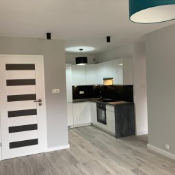 Łukasz Niedzielan - Nowoczesne wnętrze: biała kuchnia z czarnym blatem i panelami, drewnopodobna podłoga, białe drzwi z czarnymi pasami, minimalistyczne oświetlenie.