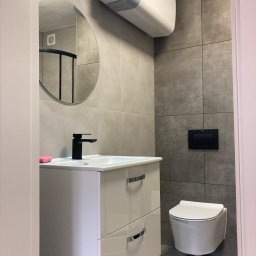 Łukasz Niedzielan - Nowoczesna łazienka z szarymi płytkami, białą szafką pod umywalką z czarną armaturą i podwieszaną toaletą. Okrągłe lustro i bojler nadają wnętrzu minimalistyczny charakter.