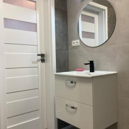 Łukasz Niedzielan - Nowoczesna łazienka z szarą płytką, białą szafką pod umywalką z czarną baterią i okrągłym lustrem. Białe drzwi z poziomymi panelami. Minimalistyczny design.