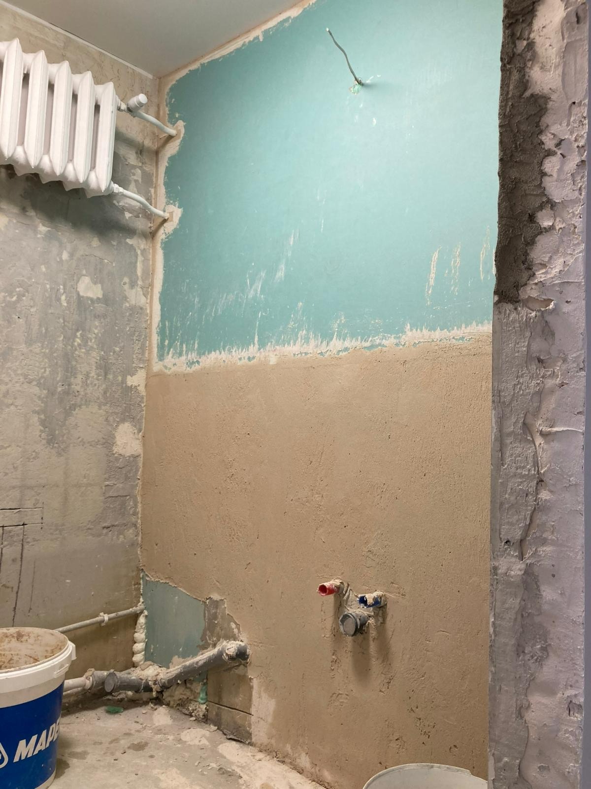 Ściana łazienki w trakcie prac: częściowo położona gładź, widoczne rury, grzejnik i surowy beton przy framudze. Górna część ściany pomalowana na niebiesko.