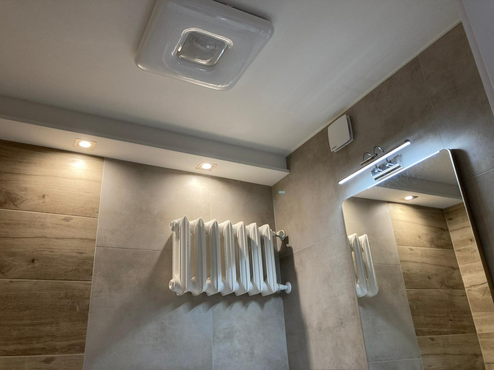 Wykończona łazienka z grzejnikiem retro, lustrem z oświetleniem LED i płytkami imitującymi drewno. Widoczne oświetlenie sufitowe.