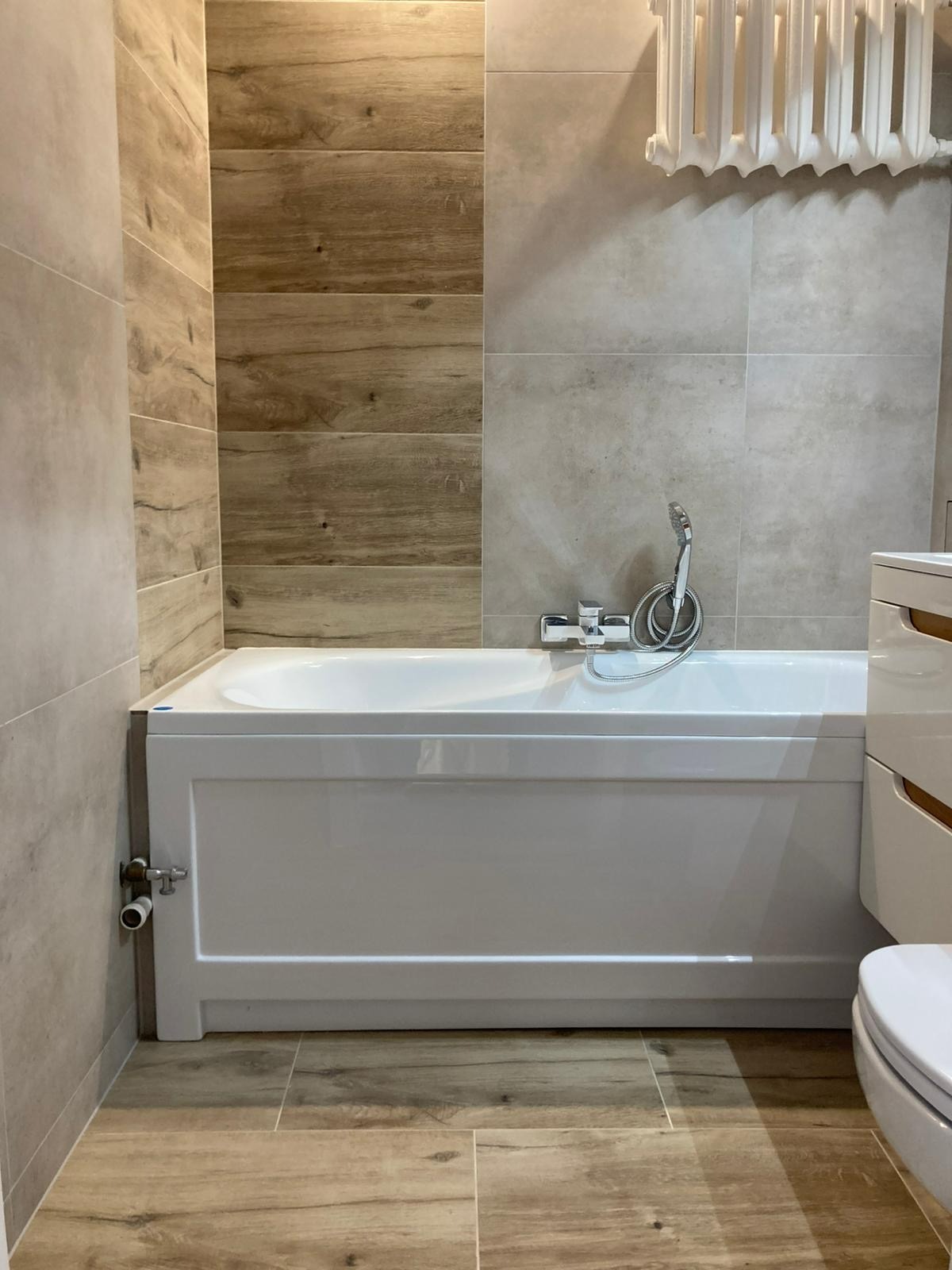 Wykończona łazienka z wanną obudowaną białą płytą, szare płytki na ścianach i drewnopodobne na fragmencie ściany oraz podłodze. Grzejnik żeberkowy.