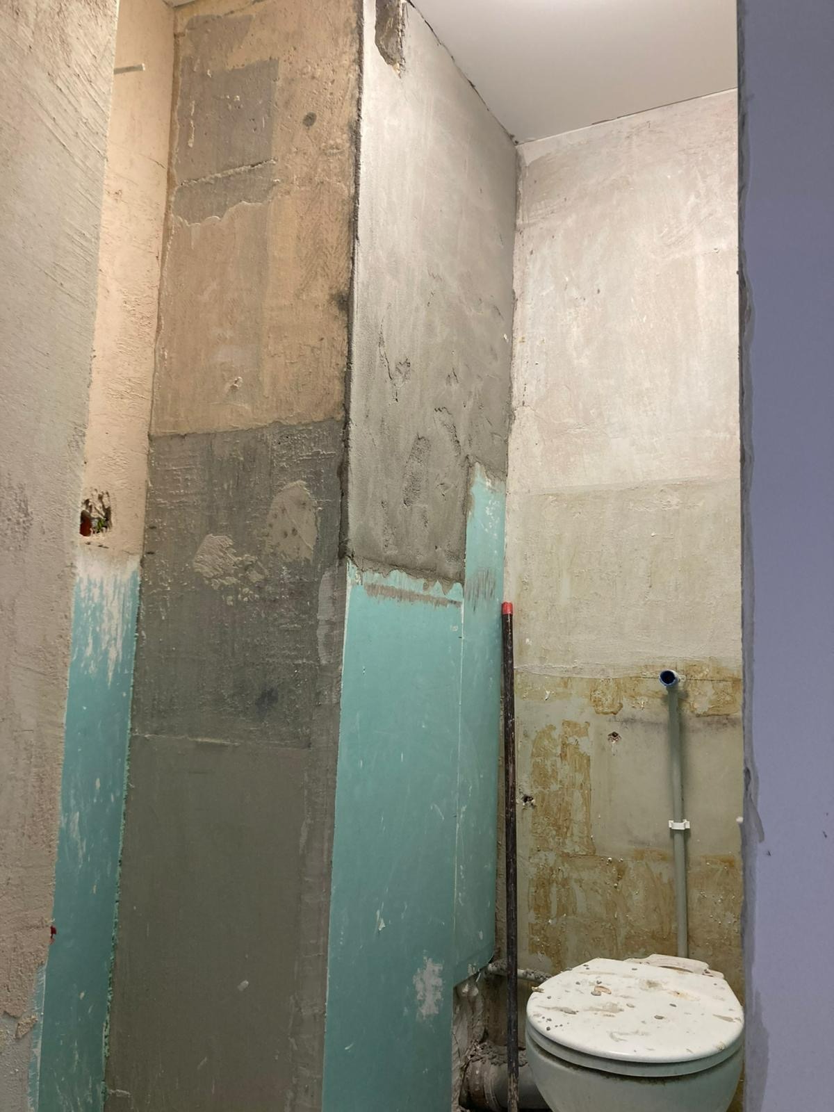 Wnętrze łazienki w trakcie remontu: surowe ściany z widocznymi warstwami tynku, fragment toalety i pionowa rura. Widoczne ślady wcześniejszych prac i przygotowanie do dalszych etapów.