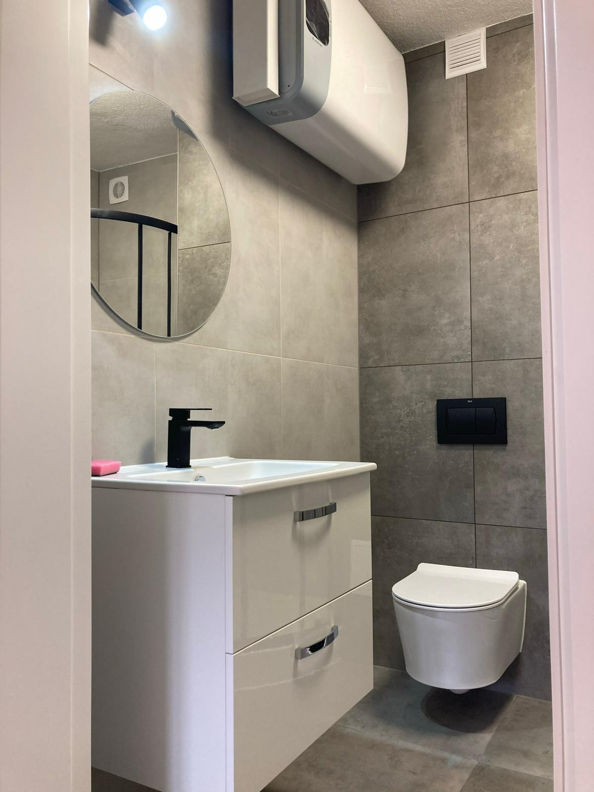 Nowoczesna łazienka z szarymi płytkami, białą szafką pod umywalką z czarną armaturą i podwieszaną toaletą. Okrągłe lustro i bojler nadają wnętrzu minimalistyczny charakter.