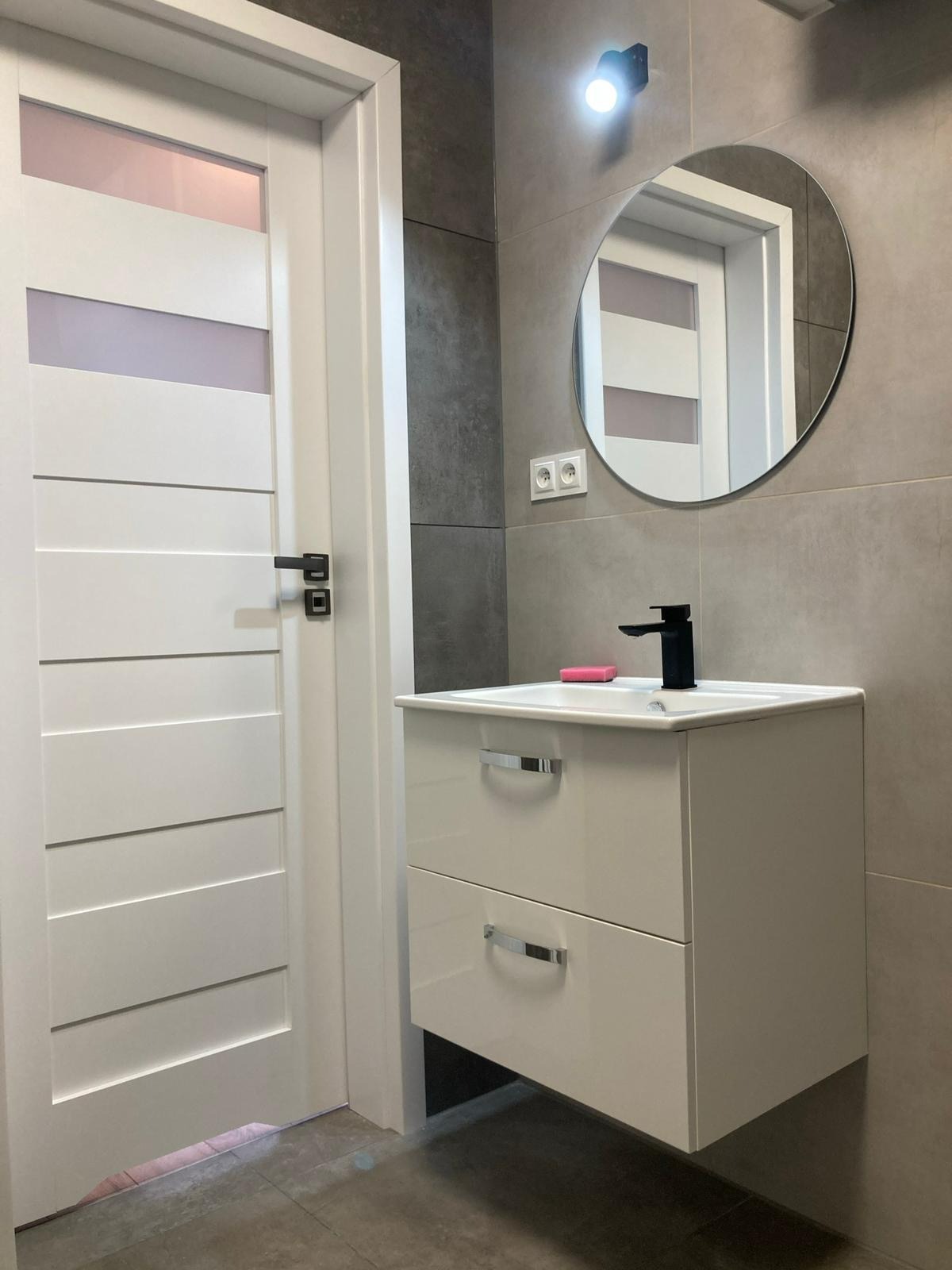 Nowoczesna łazienka z szarą płytką, białą szafką pod umywalką z czarną baterią i okrągłym lustrem. Białe drzwi z poziomymi panelami. Minimalistyczny design.