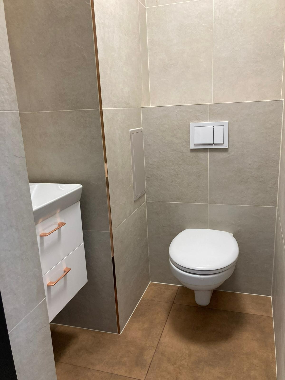 Wykończona łazienka z szarymi płytkami, białym WC i umywalką z szafką. Brązowe płytki podłogowe. Uwagę przyciągają miedziane detale wykończeniowe.