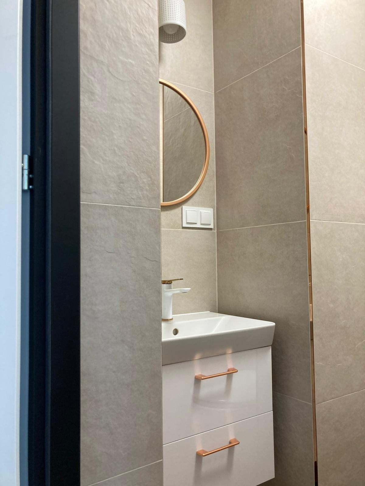 Nowoczesna łazienka z beżowymi płytkami, okrągłym lustrem w miedzianej ramie i białą szafką pod umywalką z miedzianymi uchwytami. Widok zza framugi drzwi.