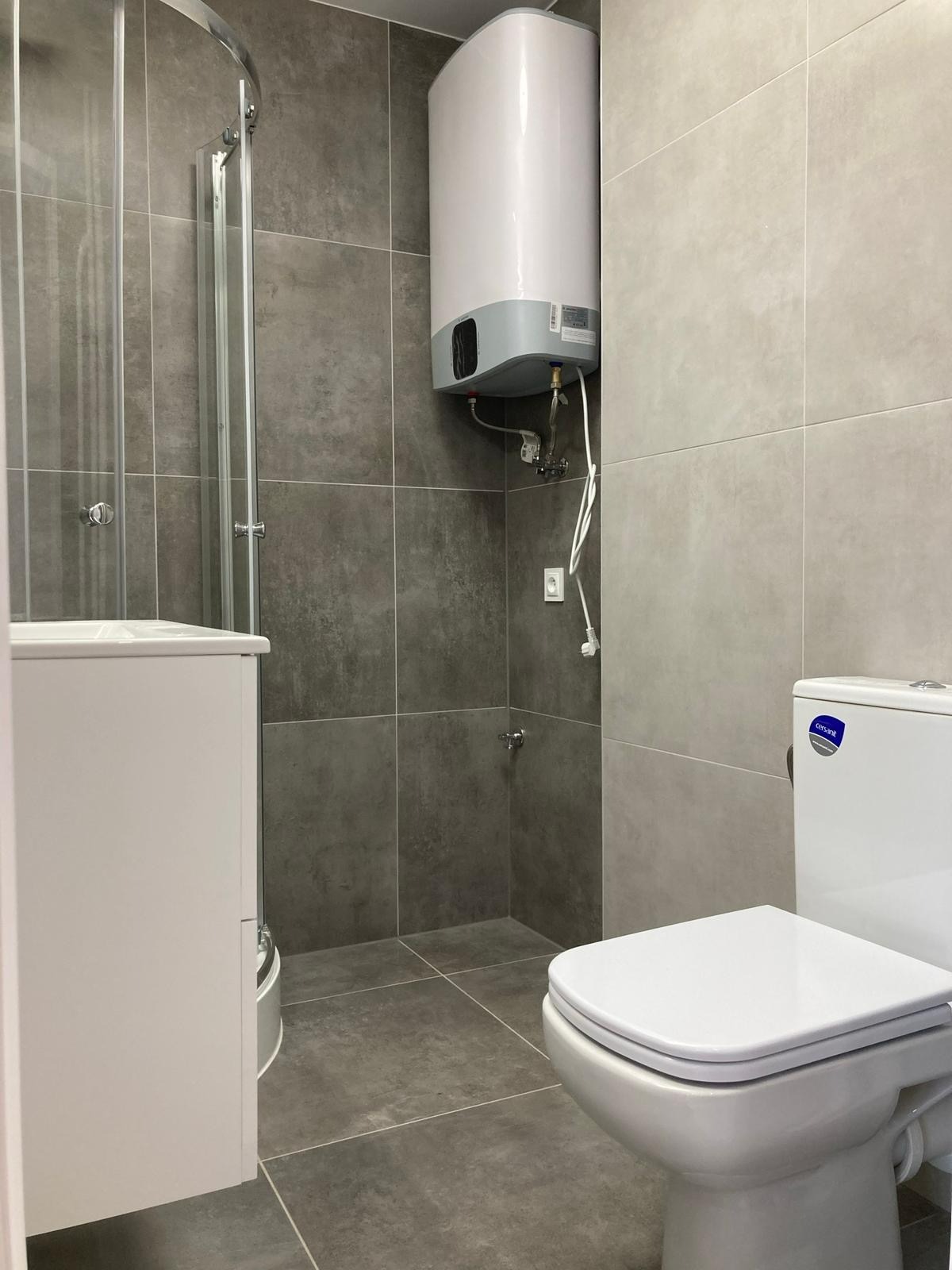 Wykończona łazienka w szarościach z kabiną prysznicową, bojlerem i toaletą. Widoczne płytki na ścianach i podłodze. Minimalistyczny design.