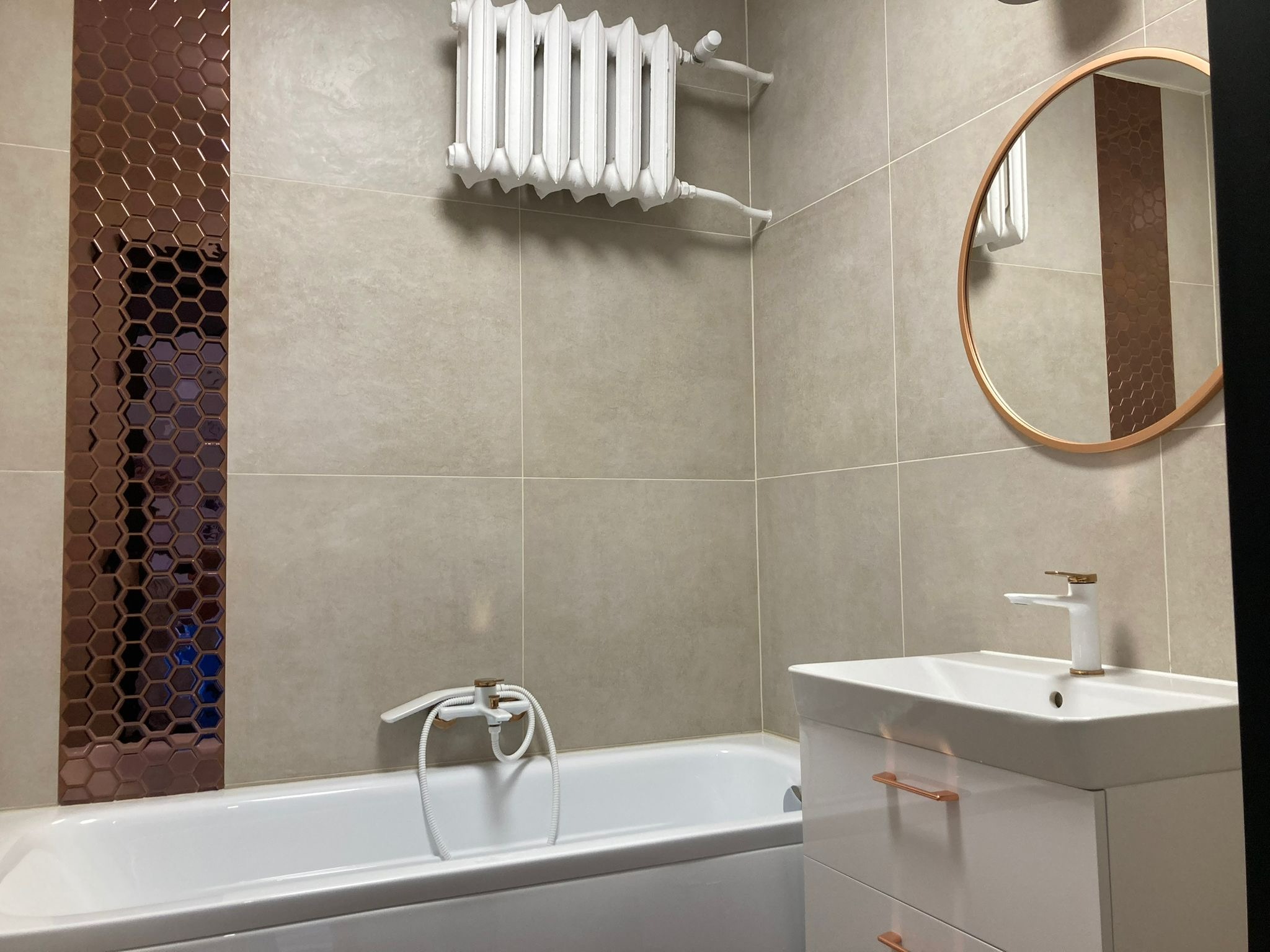 Wykończona łazienka z beżowymi płytkami, dekoracyjnym pasem mozaiki w odcieniach brązu, białą wanną i umywalką z armaturą w kolorze miedzi oraz grzejnikiem.