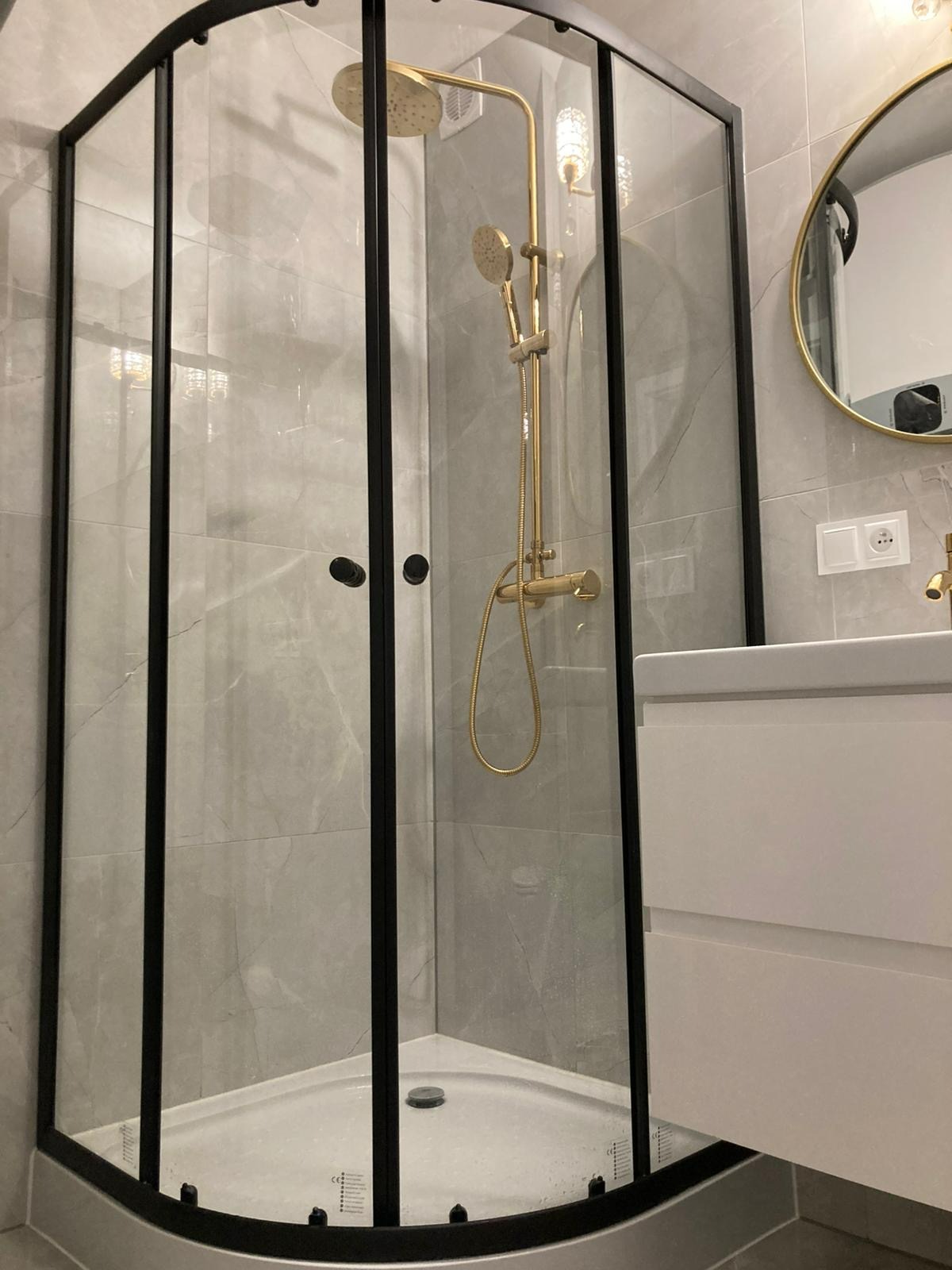 Nowoczesna kabina prysznicowa z czarnymi profilami i złotą armaturą na tle szarych płytek imitujących marmur. Umywalka z szafką w minimalistycznym stylu.
