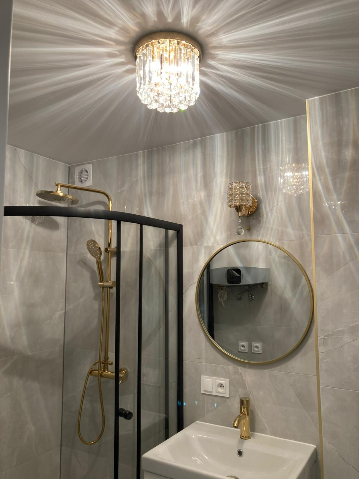 Wykończona łazienka z kabiną prysznicową typu walk-in z czarnymi profilami, złotą armaturą i okrągłym lustrem. Na suficie kryształowy żyrandol.