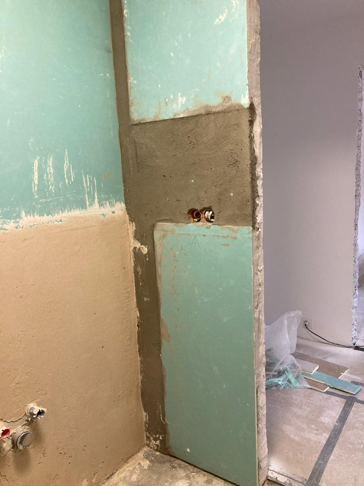 Narożnik łazienki w trakcie remontu: fragment ściany z płytą karton-gips, widoczne rury instalacji wodnej, surowy tynk. Etap prac przygotowawczych.