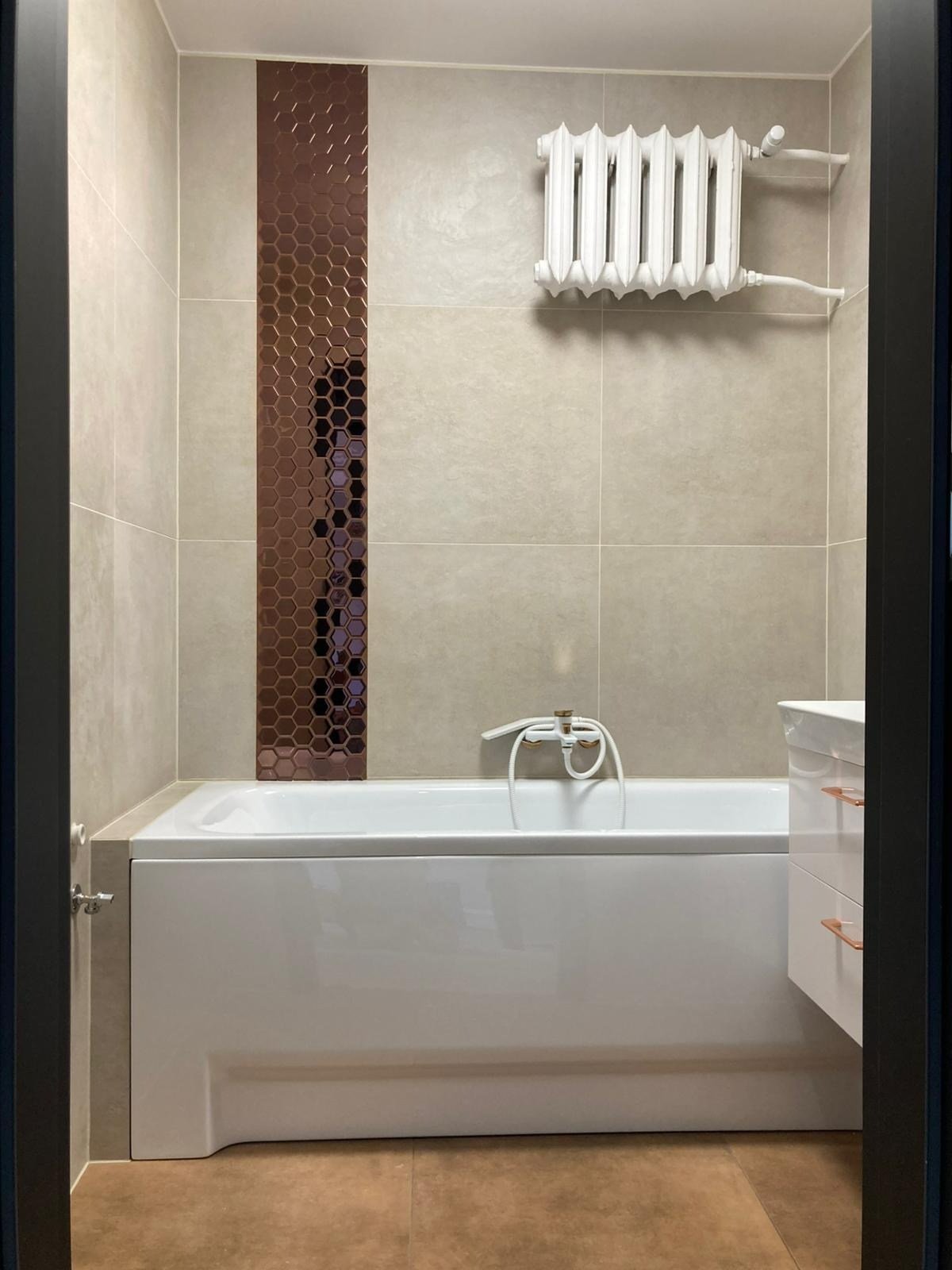 Wykończona łazienka z beżowymi płytkami, dekoracyjnym pasem mozaiki w odcieniach brązu, białą wanną i grzejnikiem żeberkowym. Widok zza framugi drzwi.