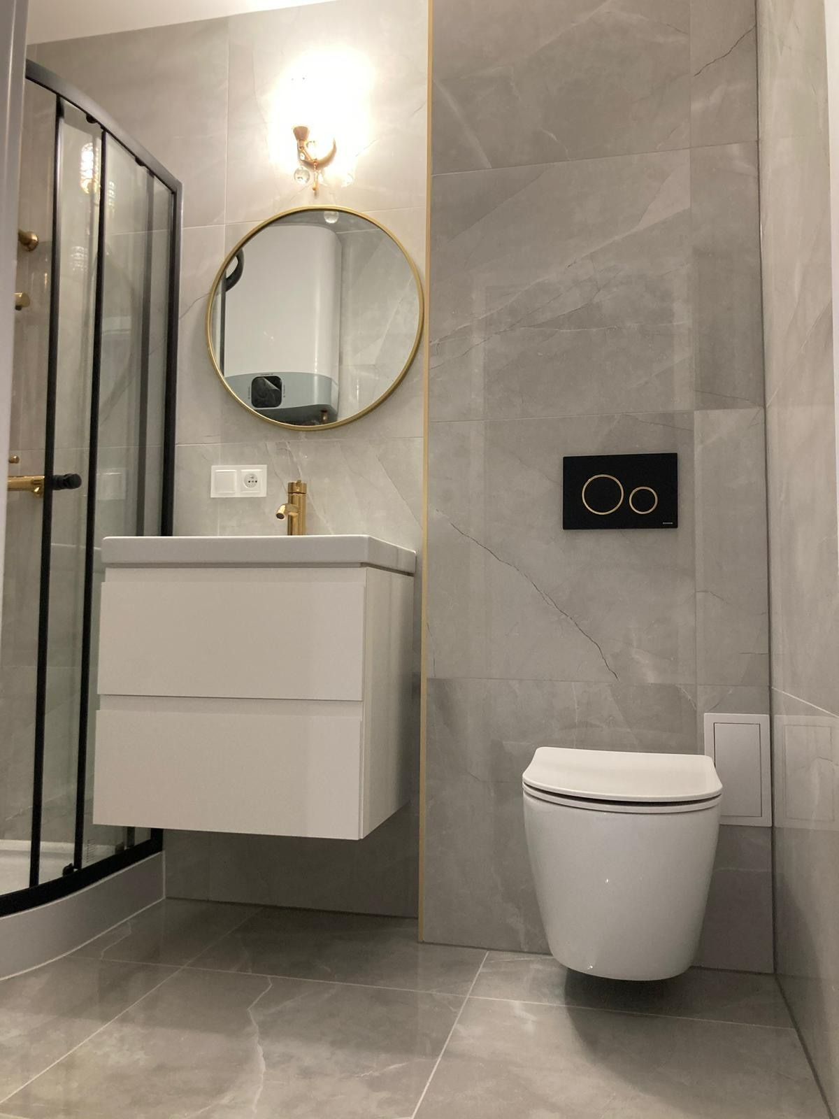 Elegancka łazienka w szarościach z kabiną prysznicową, wiszącą szafką z umywalką i toaletą. Złote akcenty: rama lustra, bateria. Płytki imitujące marmur.