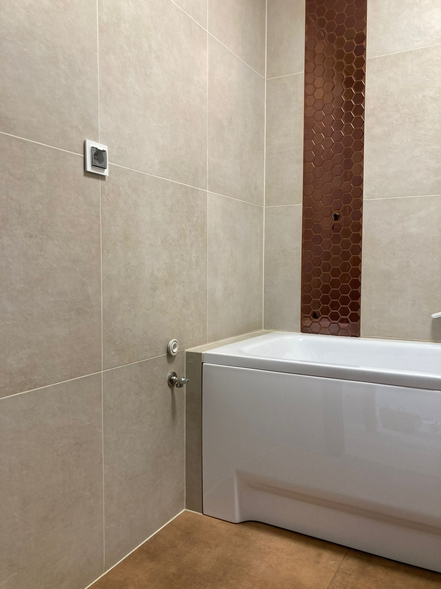 Beżowa łazienka z wanną i dekoracyjnym pasem brązowych płytek heksagonalnych. Widoczny fragment podłogi w kolorze terakoty i biały włącznik światła.
