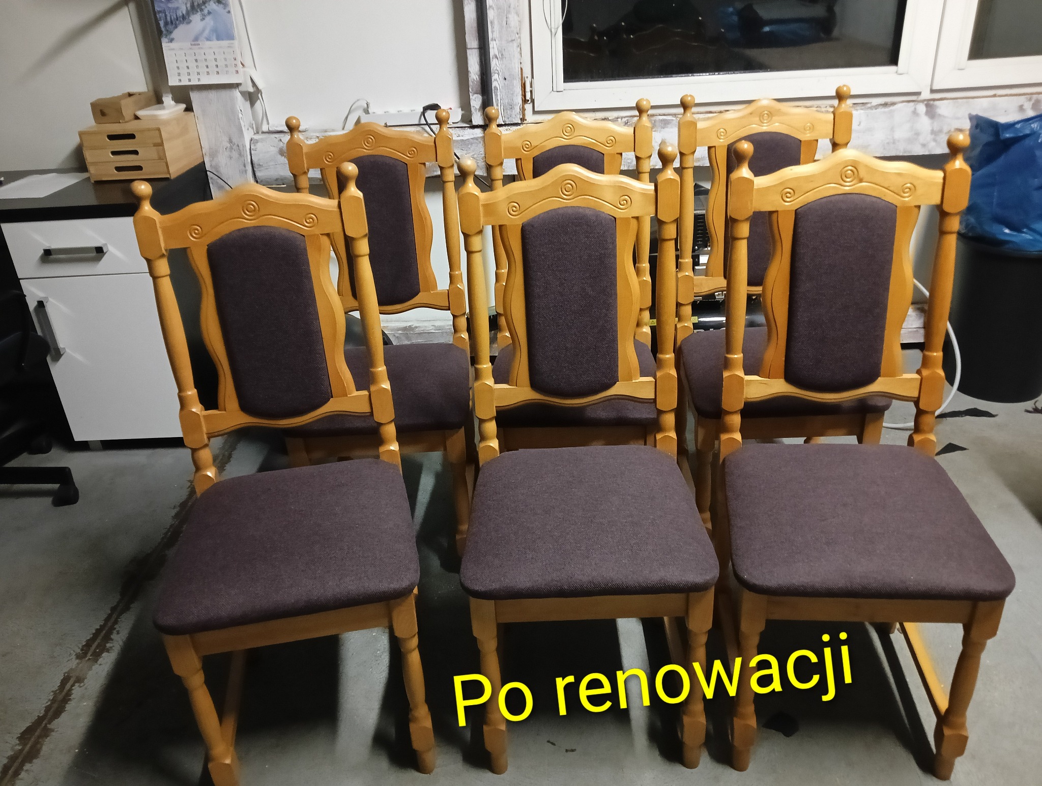 Sześć odnowionych krzeseł z drewnianą ramą i ciemnoszarym obiciem ustawionych w rzędzie, widoczne po renowacji, na tle ściany i okna.
