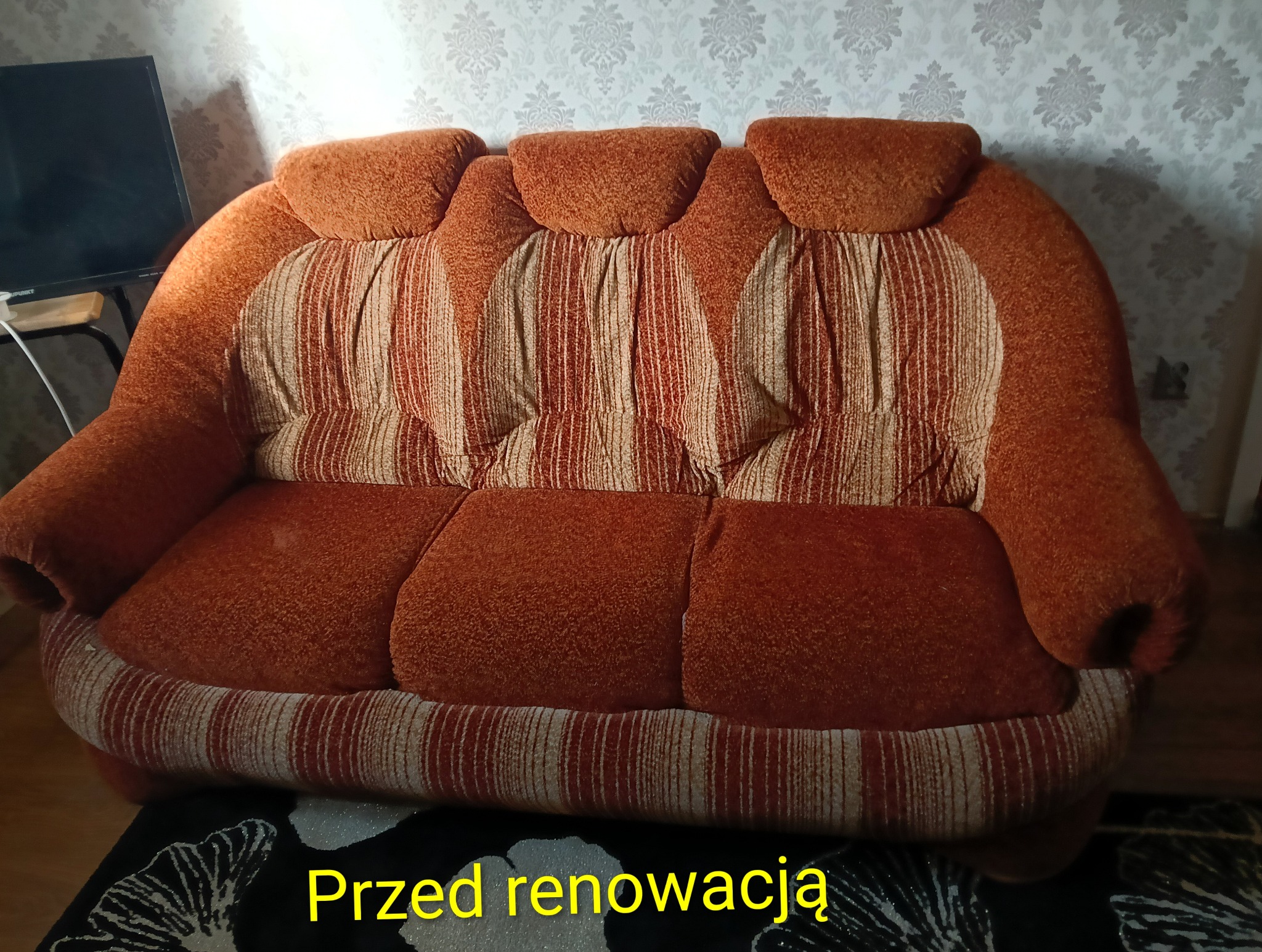 Pomarańczowa sofa z wzorem w paski, widoczna w salonie przed renowacją. Widoczne ślady użytkowania i zużycia tapicerki. Tło stanowi ściana z tapetą.