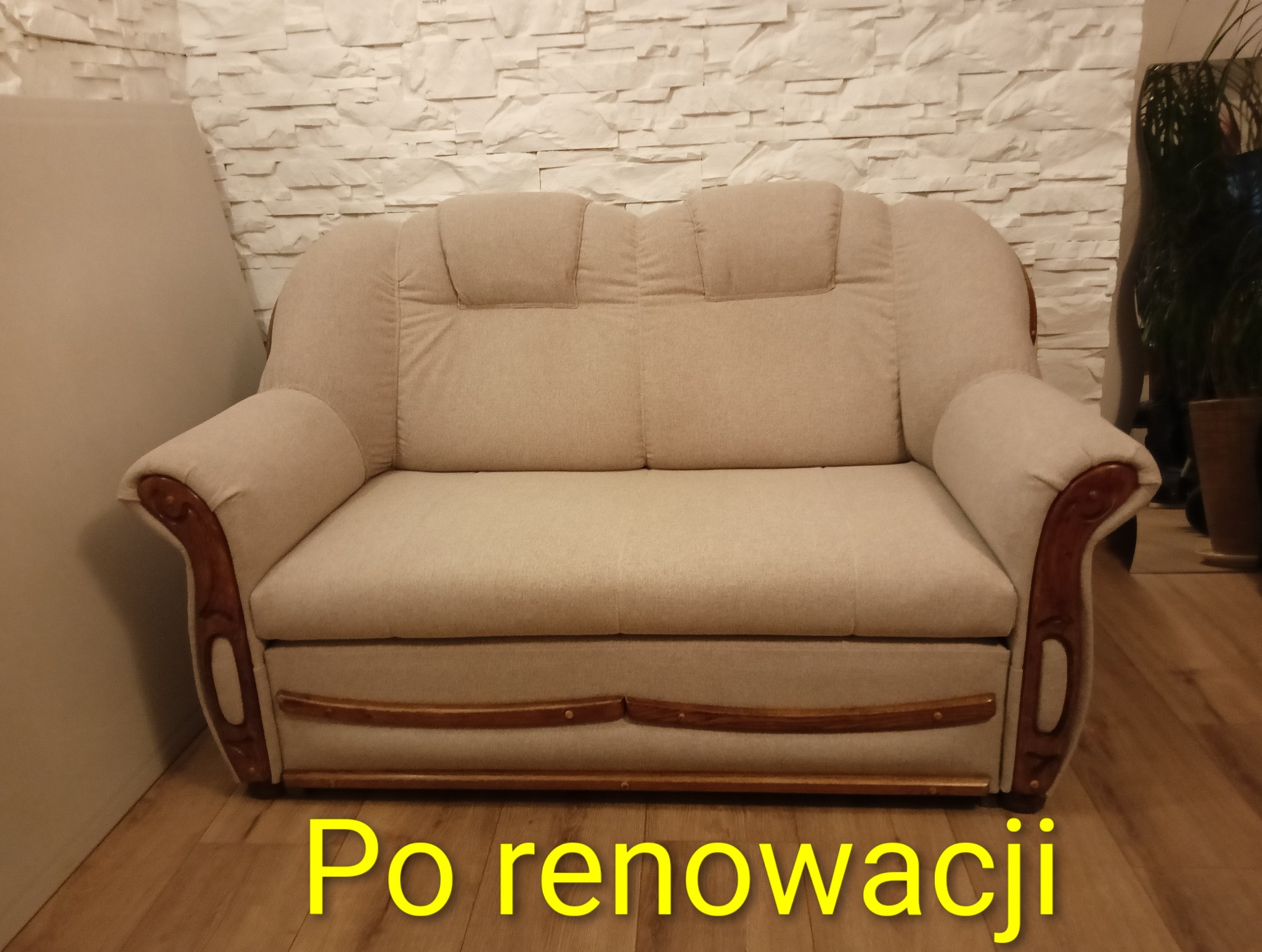 Beżowa sofa po renowacji z drewnianymi zdobieniami na tle ściany z białej cegły i drewnianej podłogi. Widoczny napis 'Po renowacji'.