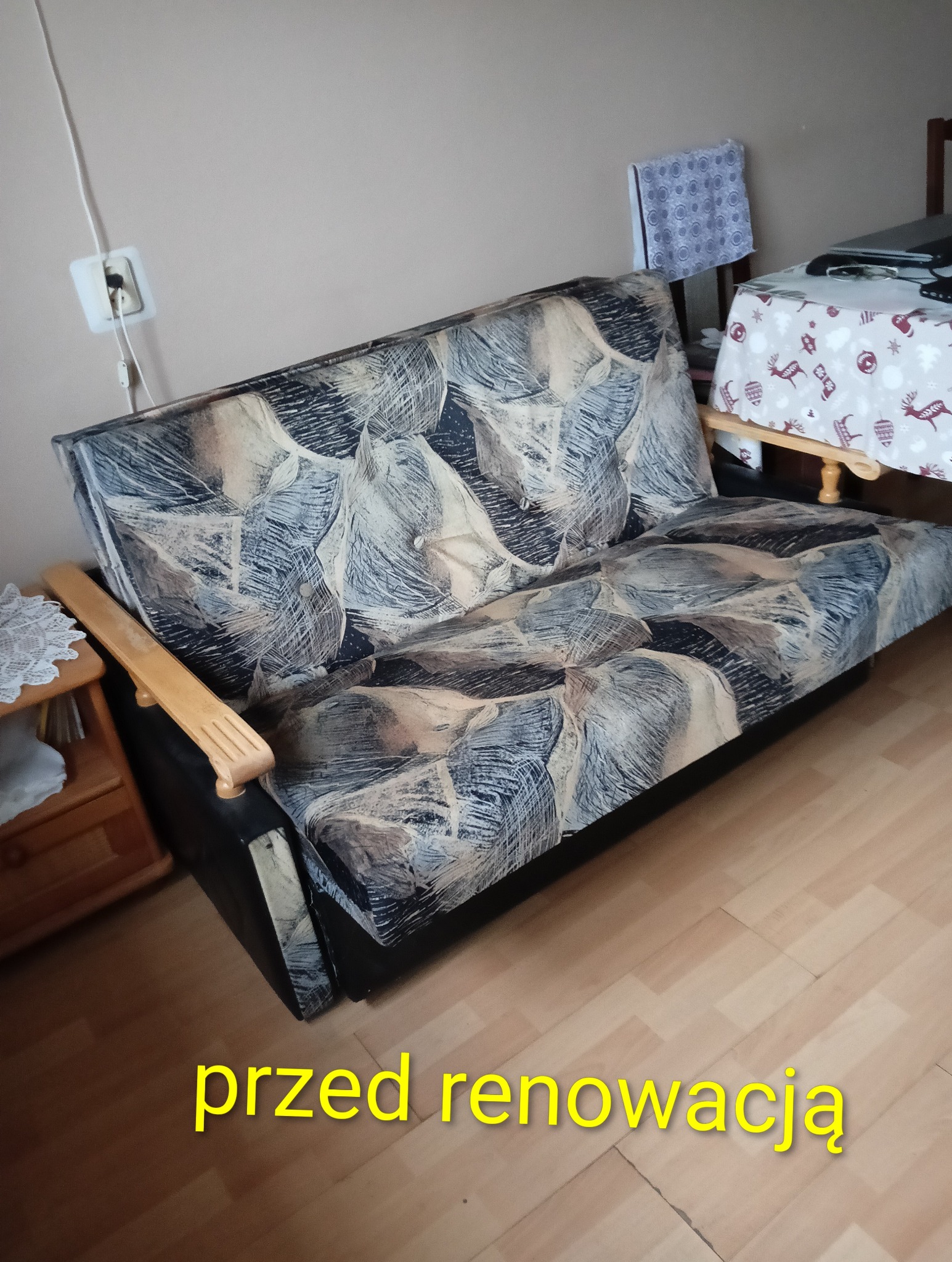 Sofa z wzorzystą tapicerką w odcieniach szarości i beżu, drewniane podłokietniki, widoczny fragment pokoju. Zdjęcie 'przed renowacją'.