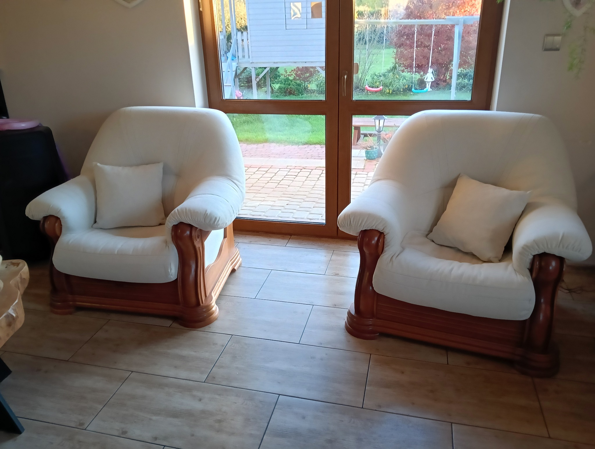 Dwa kremowe fotele z drewnianymi ramami stoją na kafelkach, z widokiem na ogród przez okno. Na każdym fotelu leży biała poduszka.