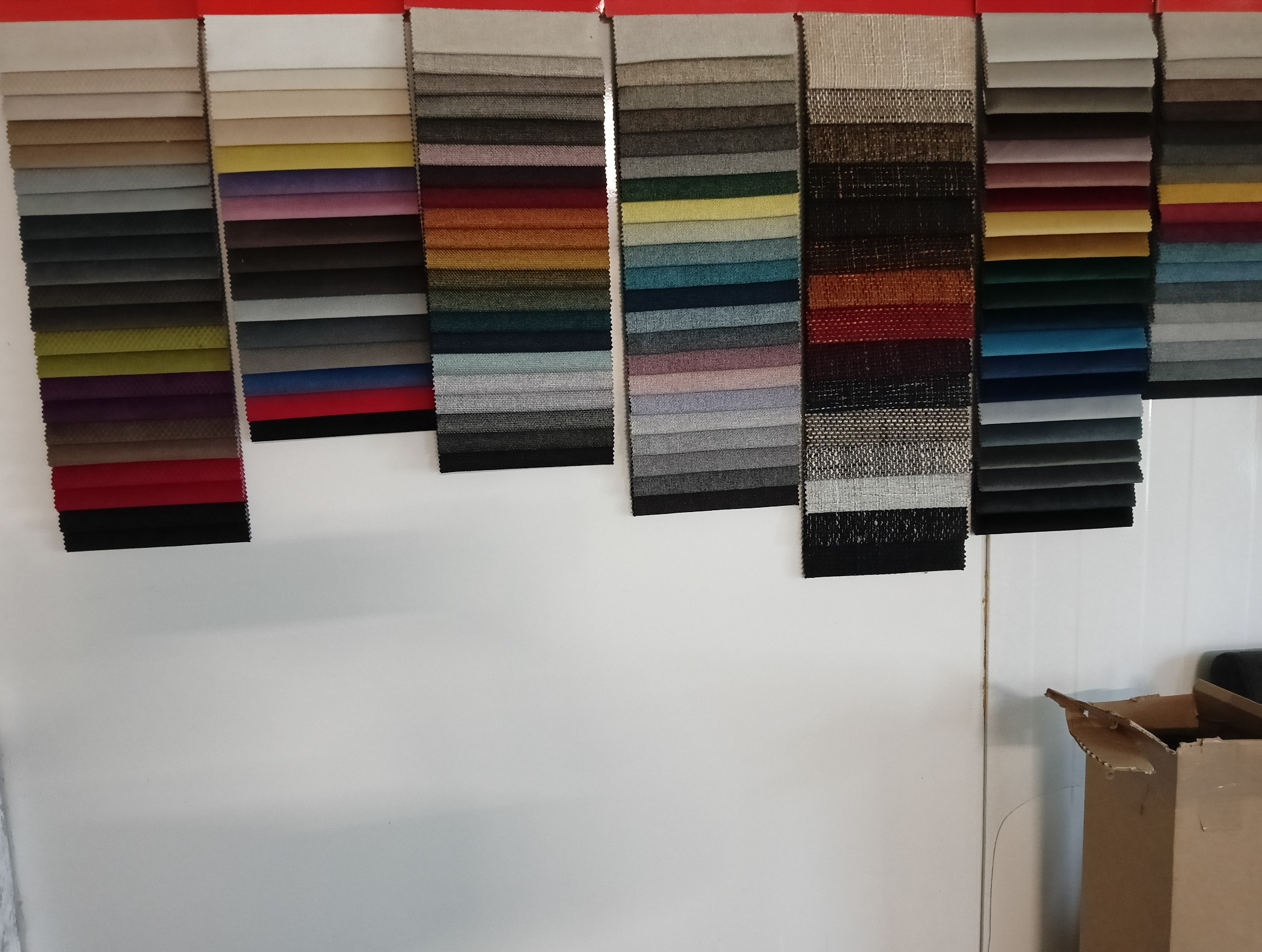 Próbki tkanin tapicerskich w różnych kolorach i fakturach, wiszące na białej ścianie. Widoczne fragmenty kartonowego pudła w prawym dolnym rogu.