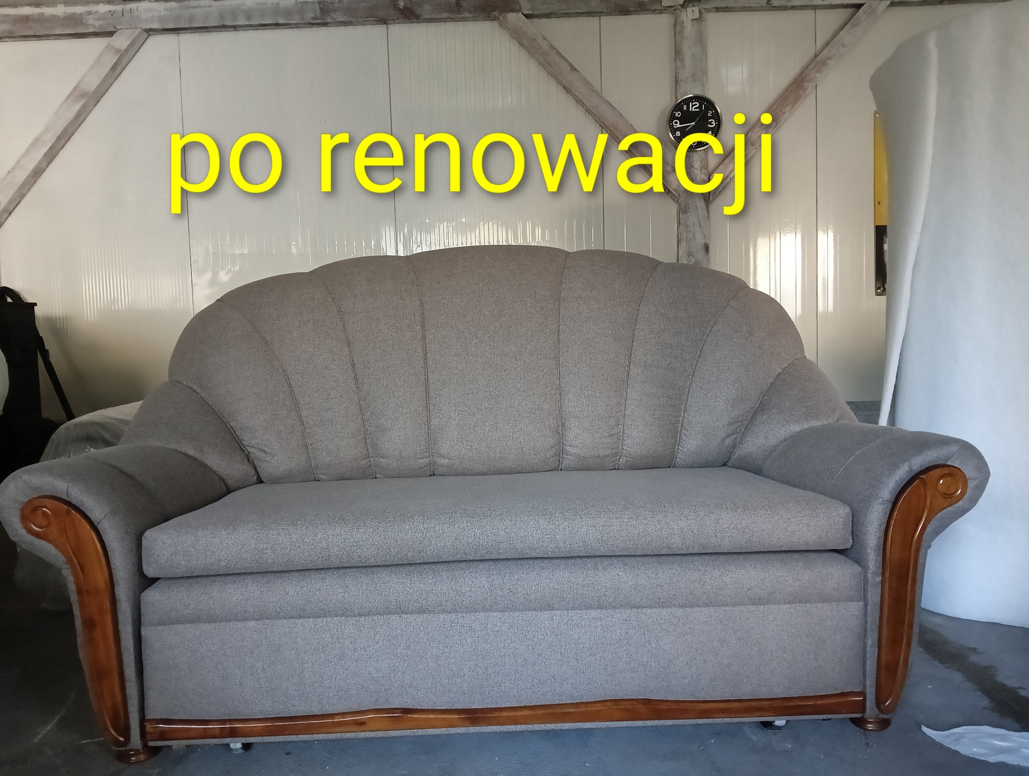 Szara sofa po renowacji z drewnianymi zdobieniami na tle białej ściany i wiszącego zegara. Widoczne detale tapicerki i odświeżony wygląd mebla.