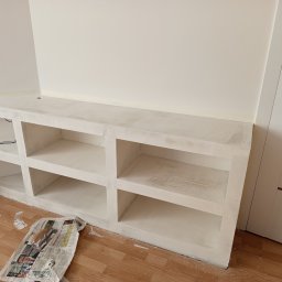 Majstereczek - Biała zabudowa meblowa z półkami wnękowymi, świeżo po malowaniu, z widocznymi śladami farby na podłodze i zabezpieczającym papierze. Widok na fragment wnętrza.