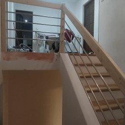 Majstereczek - Nowe, drewniane schody z metalowymi barierkami w jasnym wnętrzu. Widoczne narzędzia i elementy wykończeniowe. Ujęcie z dołu, podkreślające konstrukcję.