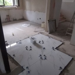 Majstereczek - Układanie białych płytek podłogowych z marmurowym wzorem, z użyciem niebieskich krzyżyków dystansowych w surowym wnętrzu remontowanego pomieszczenia.