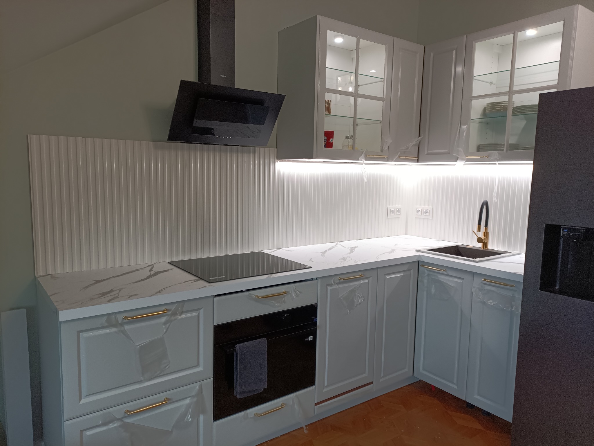 Nowoczesna, jasna kuchnia z białymi szafkami, złotymi uchwytami i marmurowym blatem. Uwagę zwraca ryflowany panel ścienny i czarny okap.