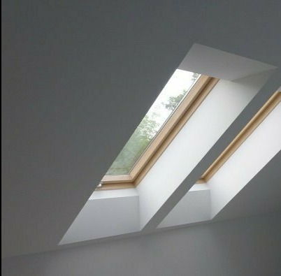 Dwa okna dachowe z drewnianą ramą, wpuszczone w białą, skośną ścianę poddasza, wpadające światło dzienne. Widok z dołu.