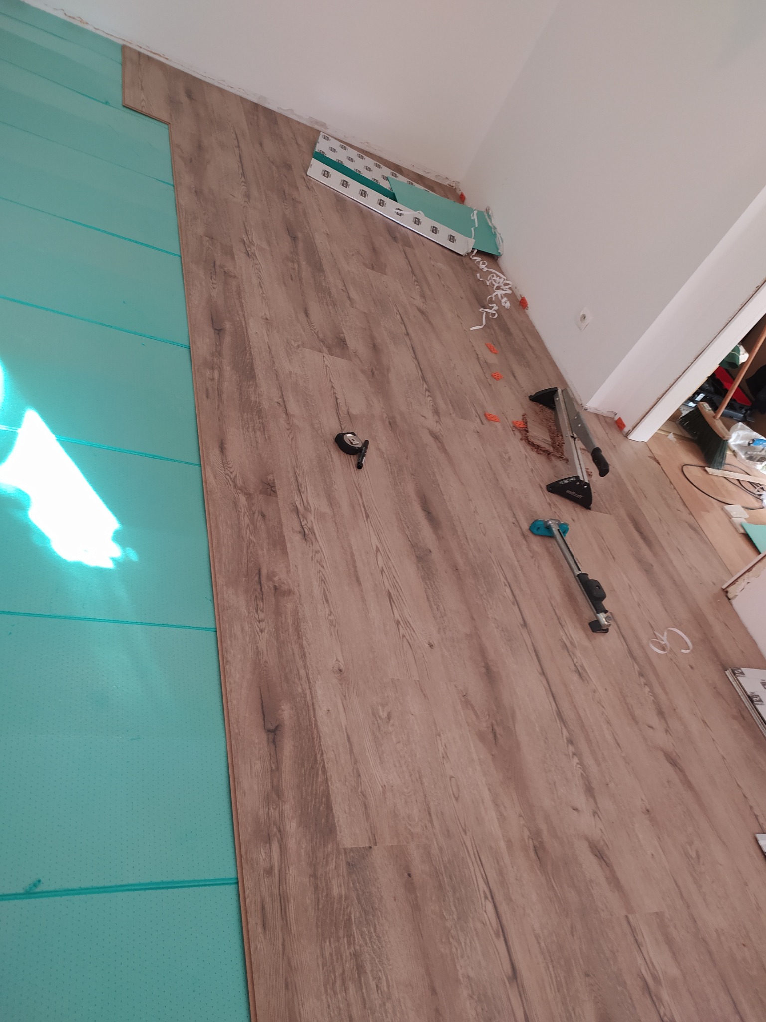 Układanie paneli podłogowych z drewnopodobnym wzorem na zielonym podkładzie. Widoczne narzędzia montażowe i elementy wykończeniowe. Ujęcie z góry, ukośne.