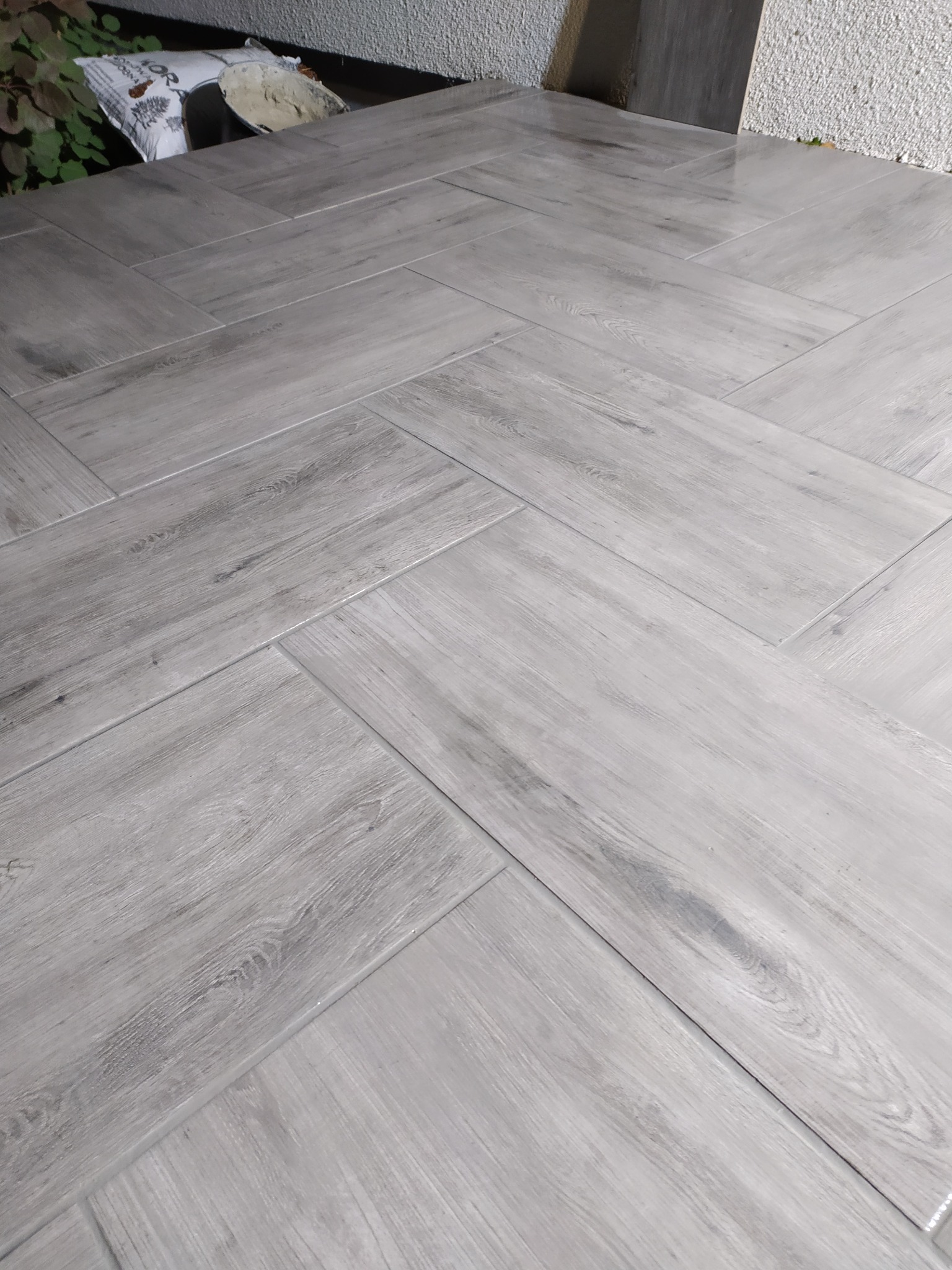 Ułożona podłoga z jasnoszarych płytek imitujących drewno w jodełkę, widoczny fragment ściany i zaprawa w worku. Minimalistyczny design podłogi.