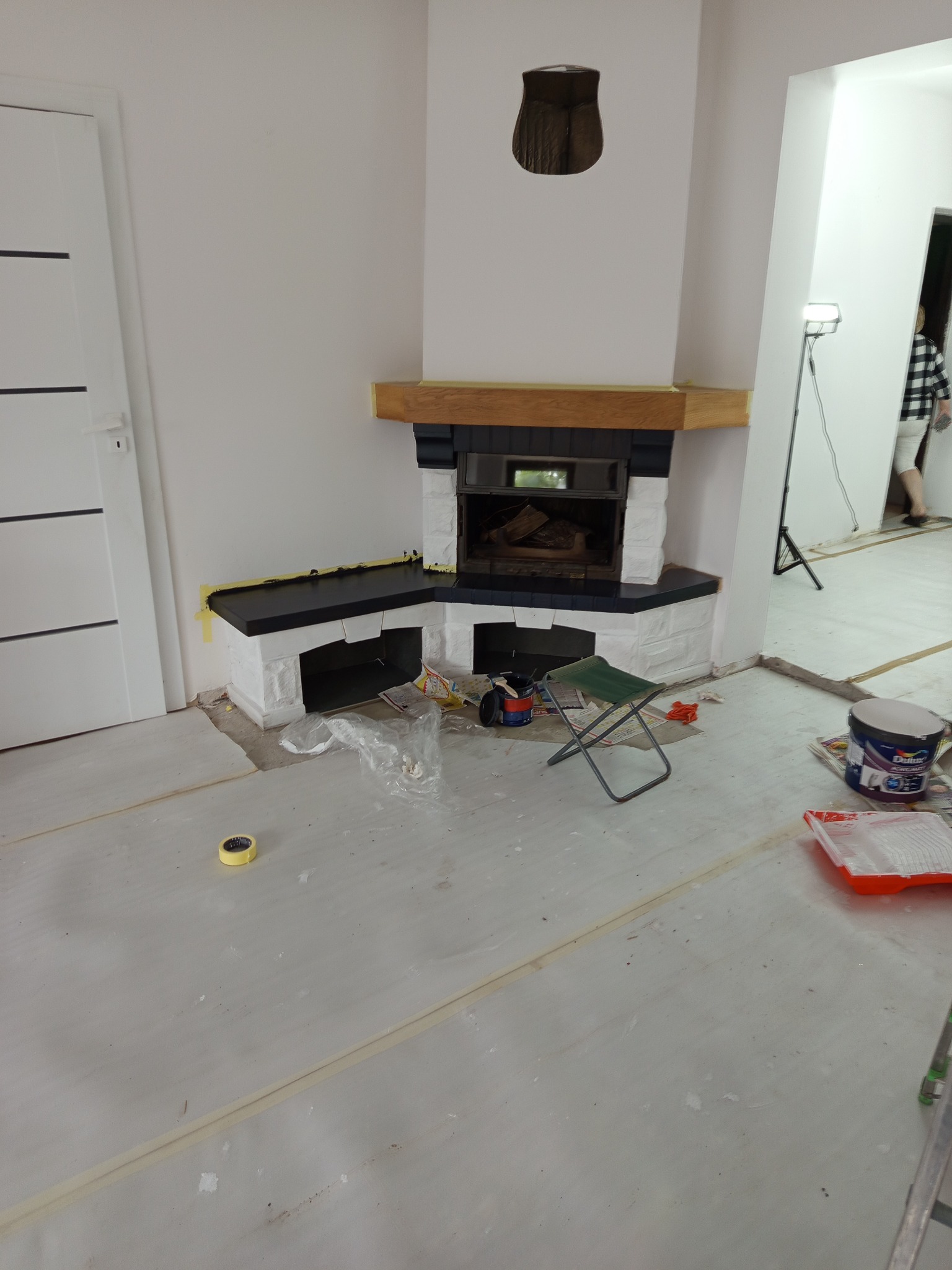 Remont pokoju z kominkiem: świeżo pomalowany kominek z drewnianą półką, zabezpieczona podłoga, narzędzia malarskie. Widok na sąsiednie pomieszczenie.