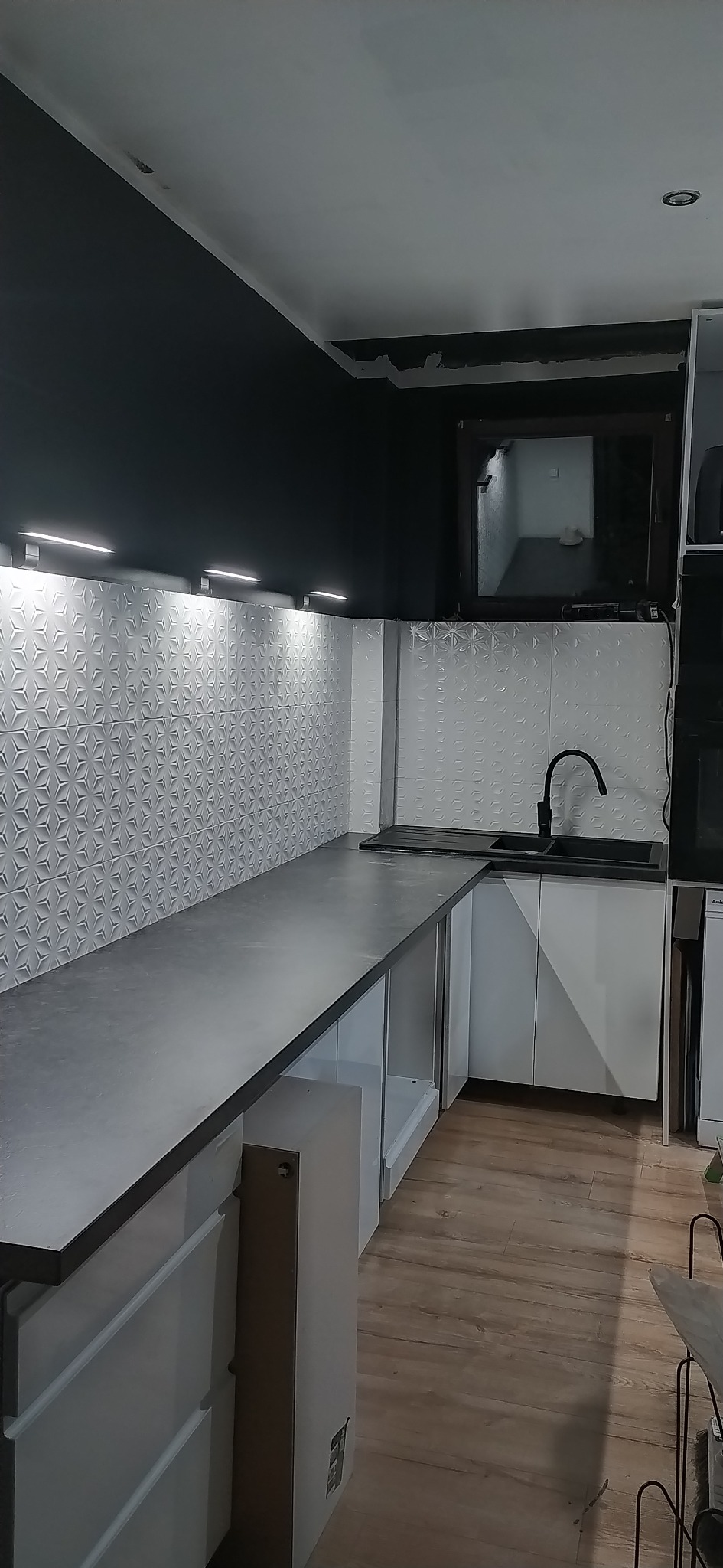 Nowoczesna kuchnia z szarym blatem, białymi szafkami i wzorzystą ścianą. Czarna armatura i oświetlenie LED podkreślają minimalistyczny design.