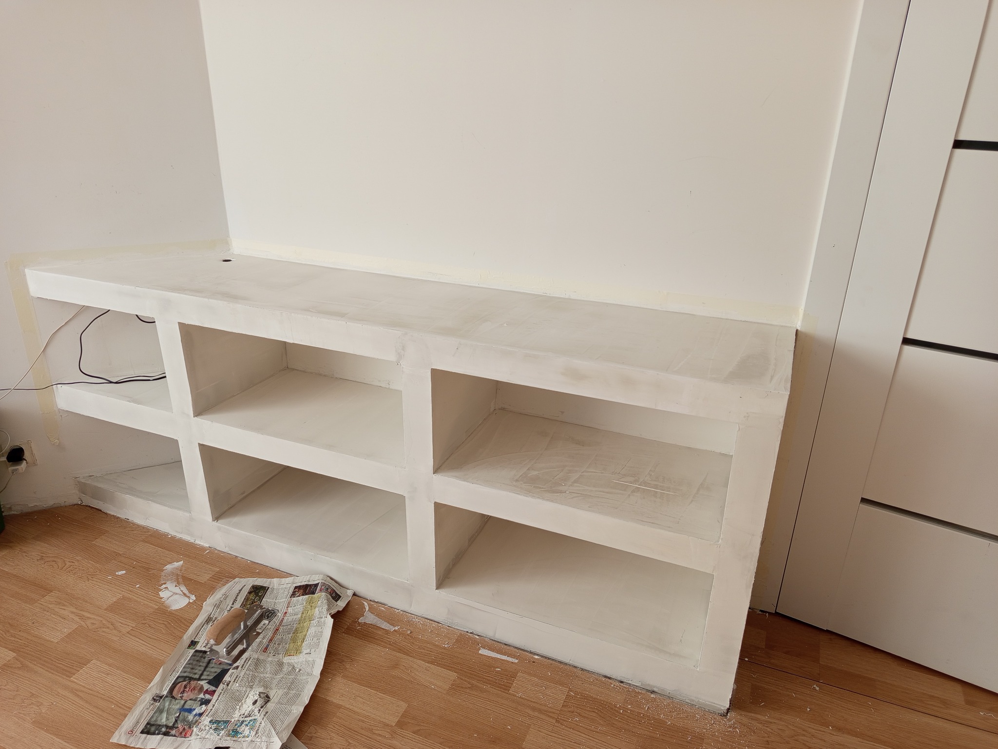 Biała zabudowa meblowa z półkami wnękowymi, świeżo po malowaniu, z widocznymi śladami farby na podłodze i zabezpieczającym papierze. Widok na fragment wnętrza.