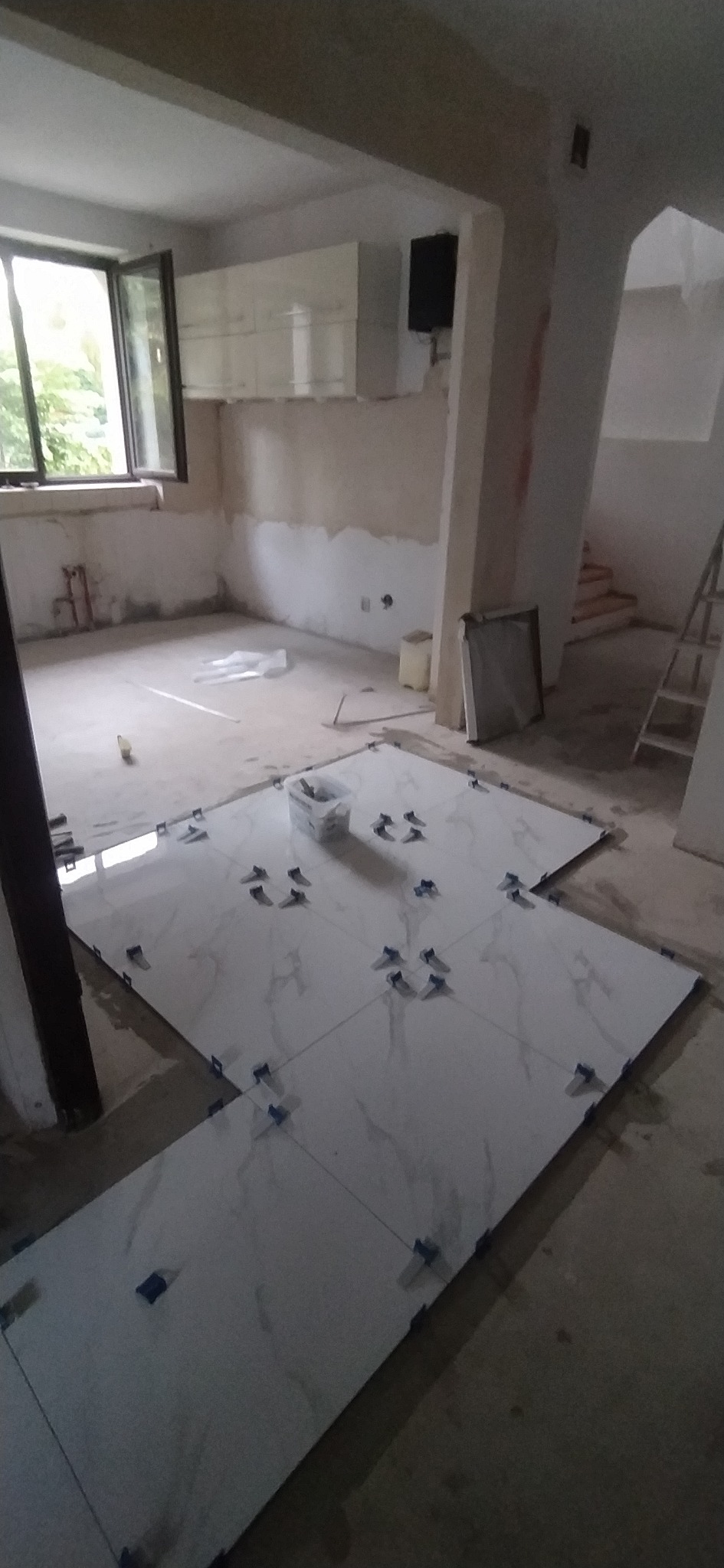 Układanie białych płytek podłogowych z marmurowym wzorem, z użyciem niebieskich krzyżyków dystansowych w surowym wnętrzu remontowanego pomieszczenia.