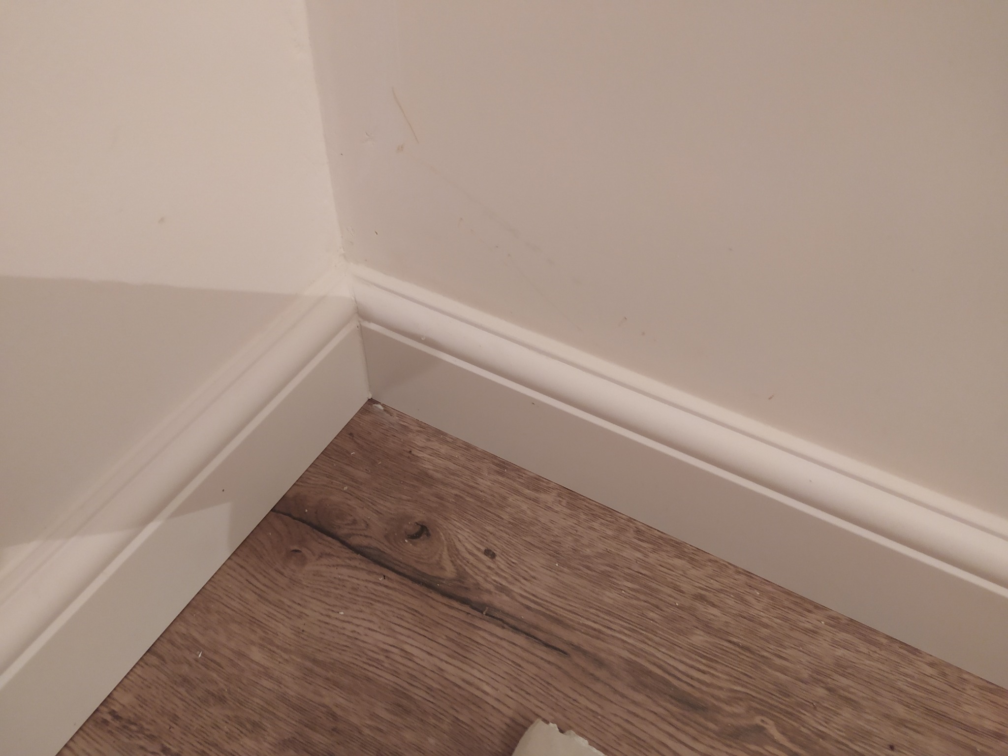 Zamknięty kadr na brązową podłogę z paneli i białe listwy przypodłogowe przy ścianie w rogu pomieszczenia. Widoczne drobne zabrudzenia na ścianie.