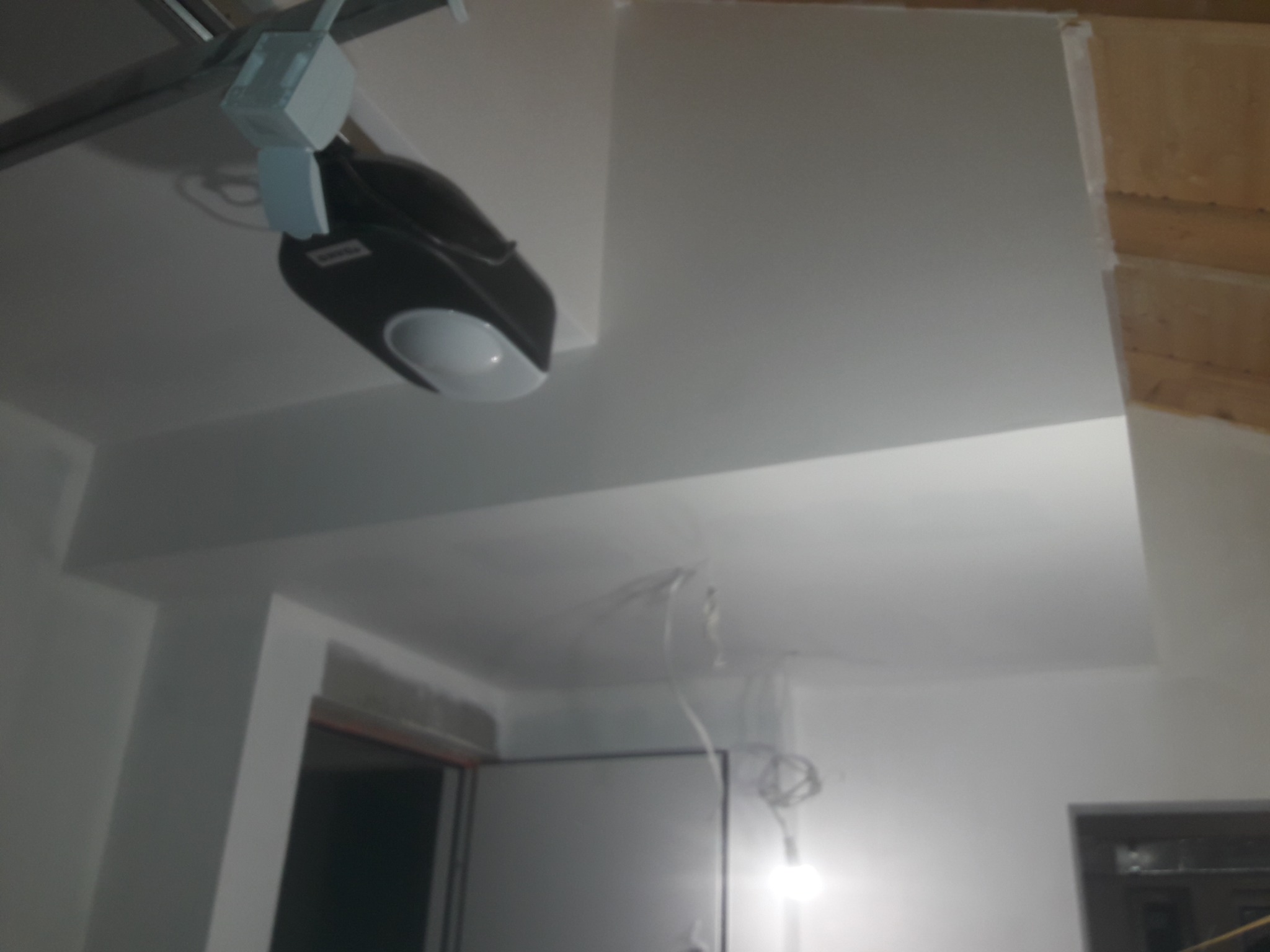 Białe, świeżo pomalowane wnętrze z widocznym mechanizmem bramy garażowej pod sufitem. Widoczne kable i fragmenty drewnianej konstrukcji.